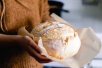 girl wearing a burn orange sweater holding a loaf of sourdough bread with parchment paper on the bottom of the loaf