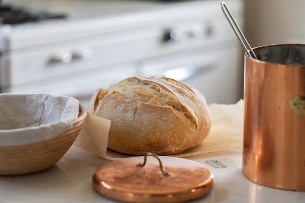 beginners loaf of sourdough bread on parchment paper on a white countertop with a copper container to the right and a banneton basket to the left