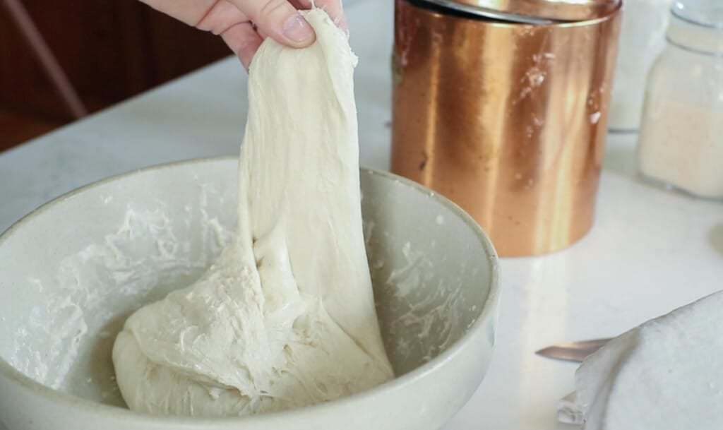 hand holding onto bread dough stretching it upwards from a stoneware bowls