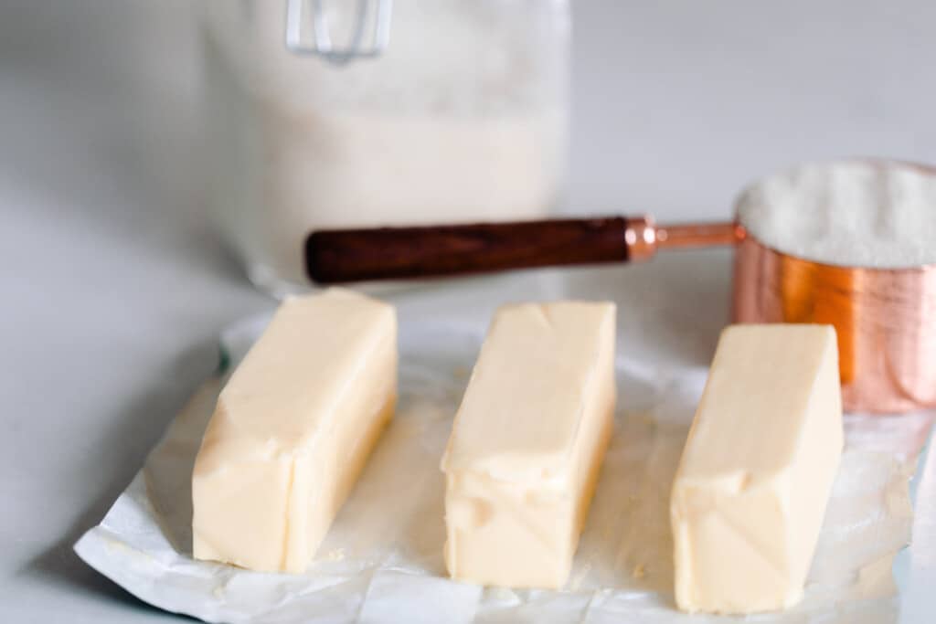 three sticks of butter with a measuring cup and jar of sourdough starter in the background