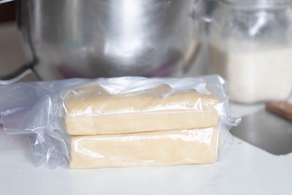 two bricks of sourdough shortbread cookie dough wrapped in plastic on a white counter top with a stainless steel bowl in the background