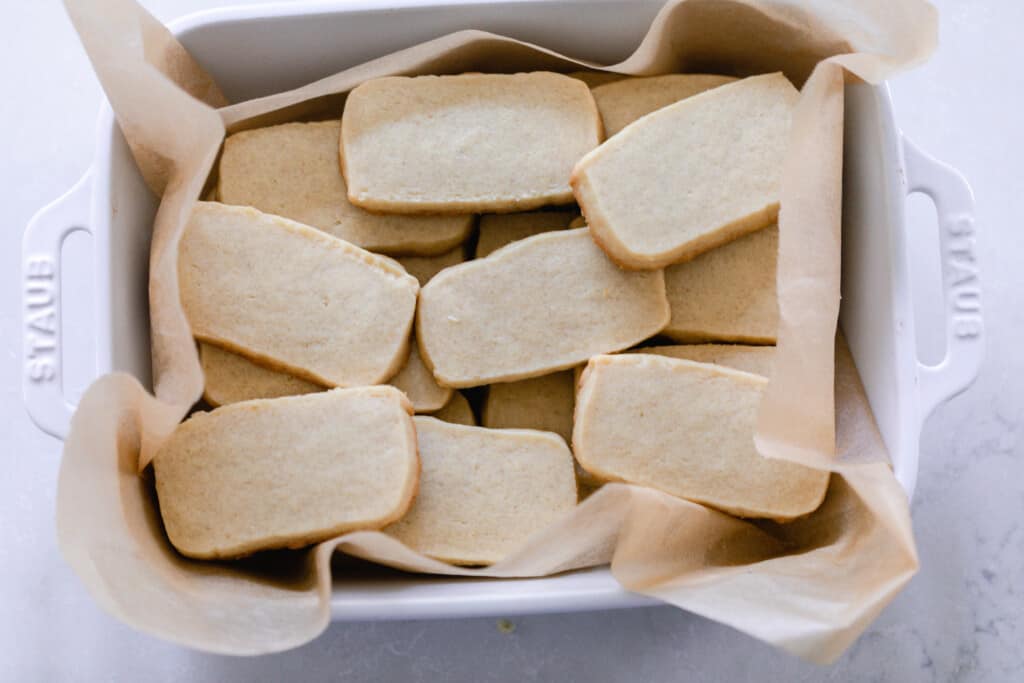 an aerial view of sourdough shortbread cookies stacked up in a white casserole dish lined with parchment paper on a white counter top
