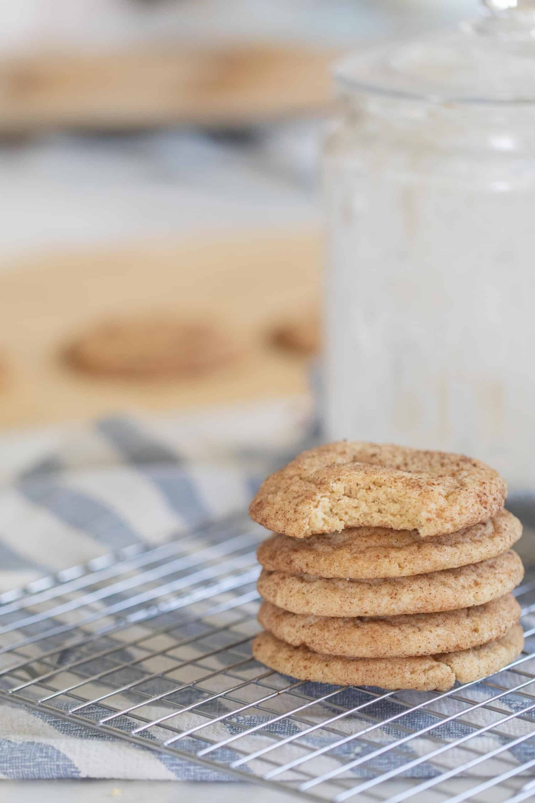 five sourdough snickerdoodle cookies stacked up on a wire rack over a white and blue stripped towel with a glass of milk behind the stack of cookies