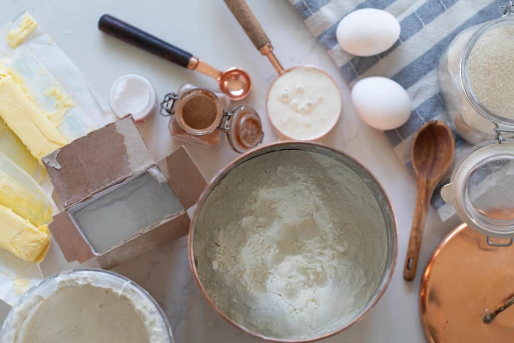 a canister of flour, measuring cups and spoons of various ingredients, a jar go sugar, sourdough starter, eggs, and butter are on a white countertop