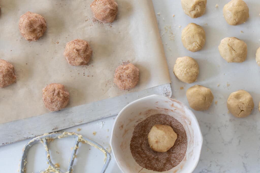 snickerdoodle cookie dough balls on a countertop. One dough ball is in a bowl with cinnamon sugar, and more dough balls with cinnamon sugar coating are on a parchment lined baking sheet