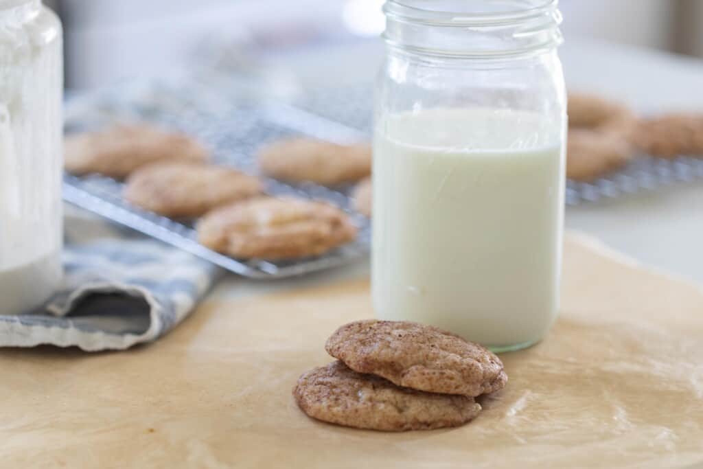 two cookies in front of a glass of milk on parchment paper. More cookies are on a cooling rack in the background