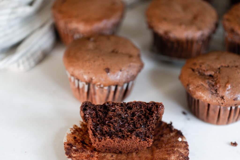 a chocolate einkorn protein muffin cut in half showing the inside texture with other muffins in the background on a white counter top