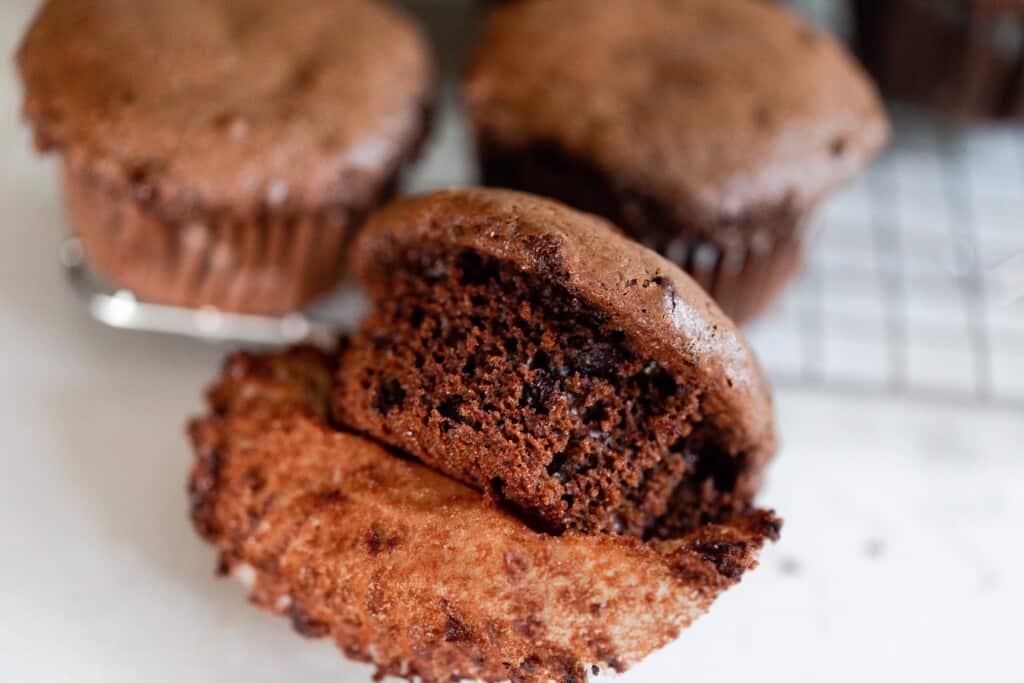 An einkorn protein muffin with the liner off to show texture and other muffins in background on cooling rack on a white counter top