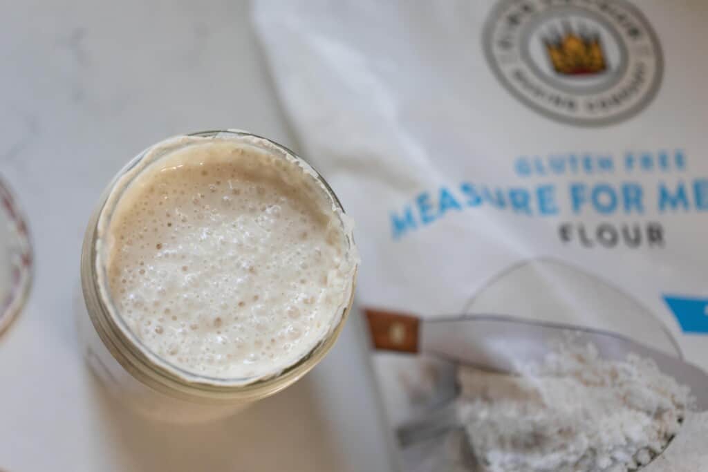 overhead photos of gluten free sourdough starter in a mason jar on a white countertop with a bag of King Arthur gluten free flour to the right