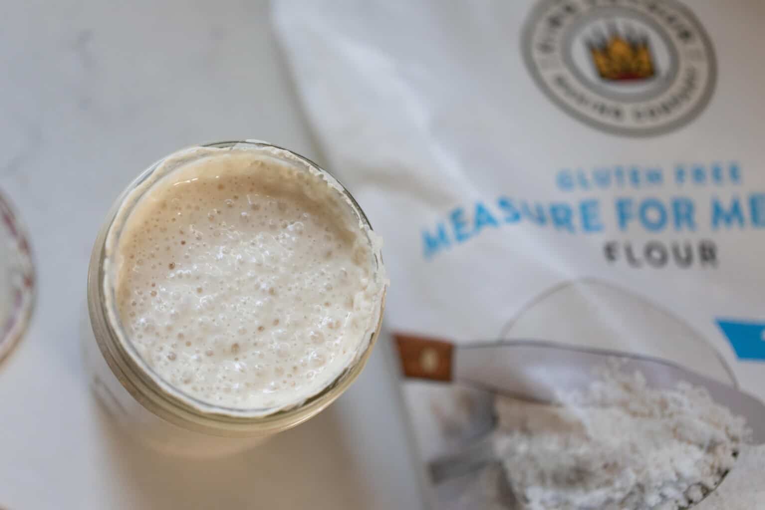 overhead photos of gluten free sourdough starter in a mason jar on a white countertop with a bag of King Arthur gluten free flour to the right