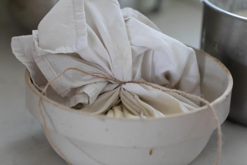 A white large bowl with a tea towel tied at the top holding the greek yogurt