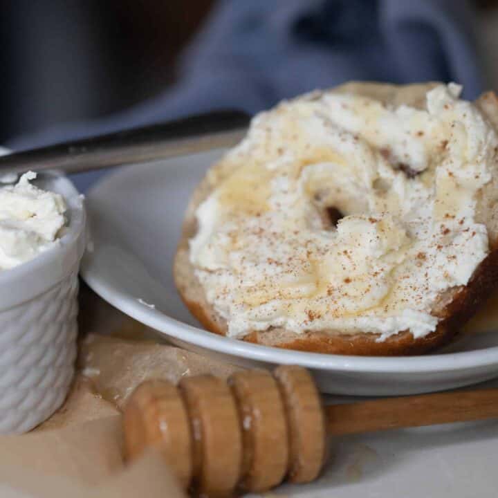 A plate with a bagel with greek yogurt cream cheese and honey with a white bowl full of cream cheese with a silver knife on top