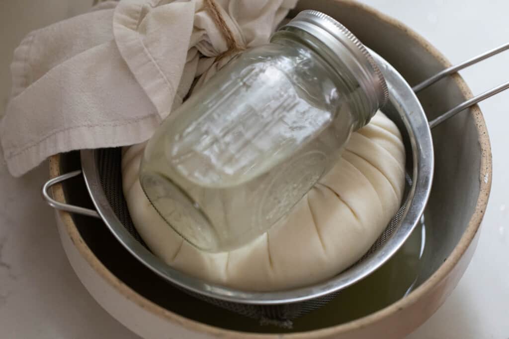 An aerial view of the greek yogurt in the tea towel over a colander and large bowl with a glass jar on top