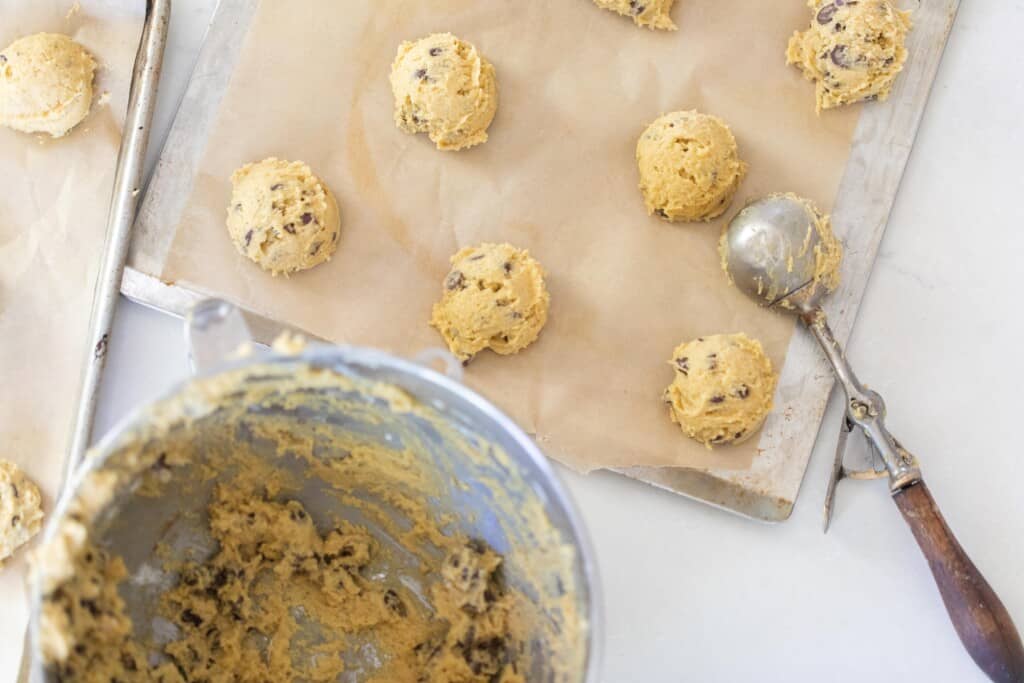 scooping chocolate chip cookie dough onto parchment lined baking sheet with a bowl of cookie dough in front of the cookie sheet