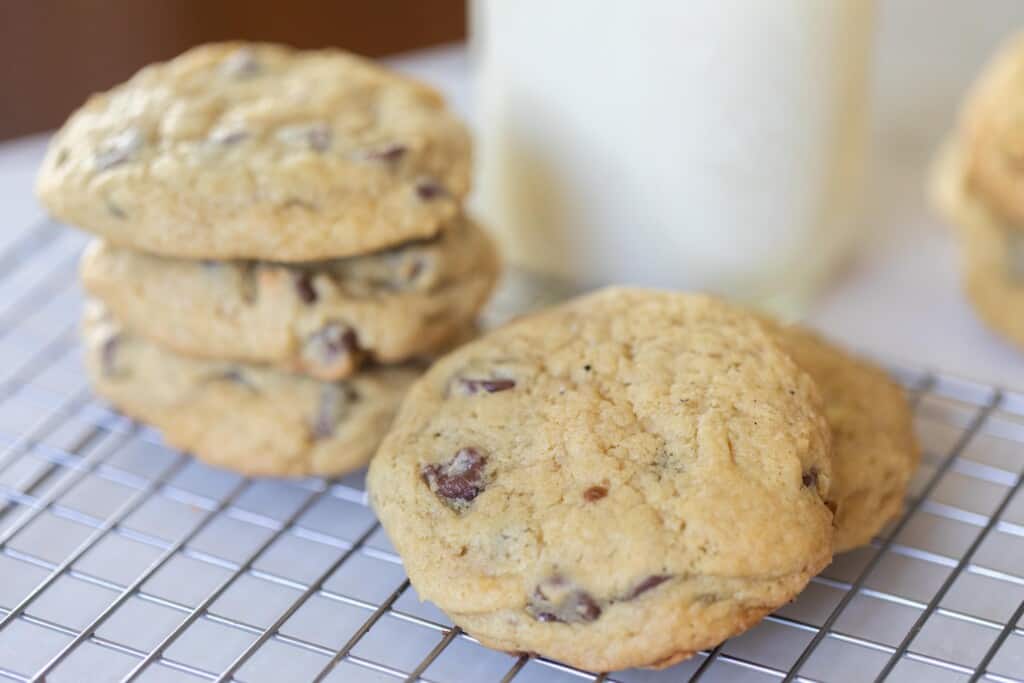 two chocolate chip cookies stacked up with another stack of cookies to the left and a glass a milk behind the cookies
