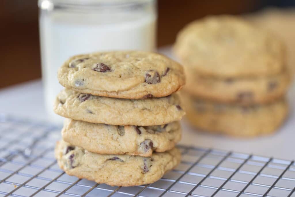 four sourdough chocolate chip cookies stack up on top of each other on a wire rack with a glass of milk and more cookies in the background