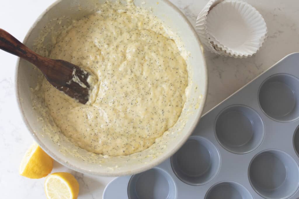 long fermented sourdough lemon poppyseed muffin batter in a large bowl with a wooden spatula with a muffin pan to the right and sliced lemons to the left