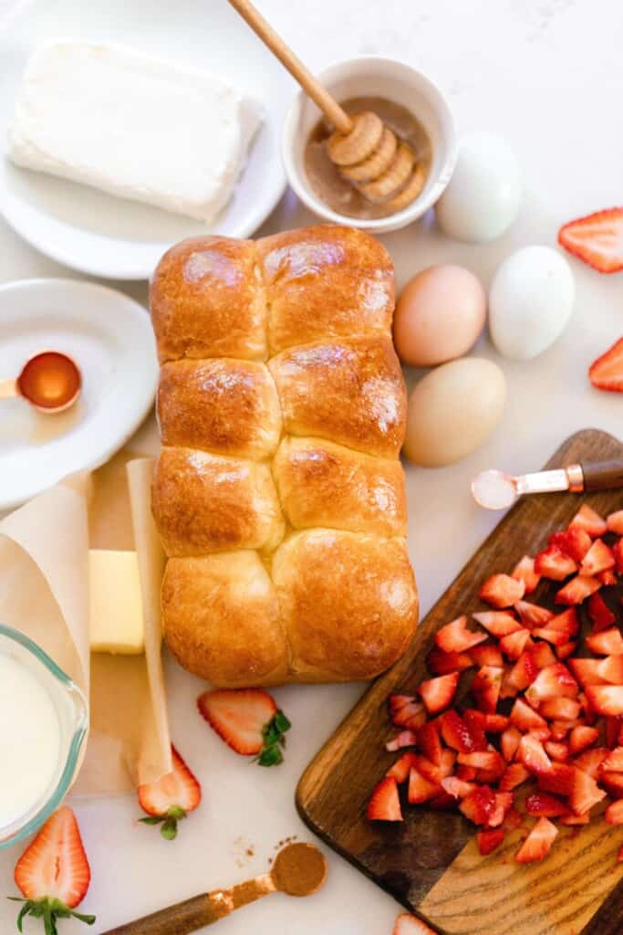 a loaf of brioche on a countertop surrounded by eggs, honey, cream cheese, and chopped strawberries on a cutting board