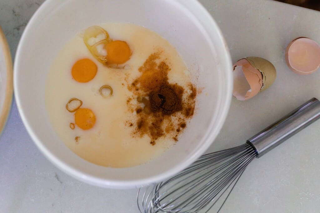 eggs, milk, vanilla, and cinnamon in a white bowl with a whisk and broken egg shells to the right