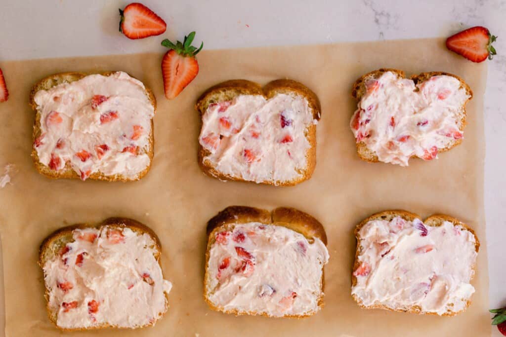 overhead photo of six slices of brioche bread covered in a strawberry cream cheese filling on parchment paper with fresh strawberries scattered around