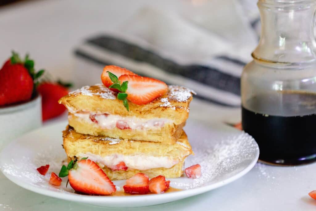 strawberry stuffed French toast stacked on a white plate and topped with strawberries. A jar of maple syrup, a blue and white towel and fresh strawberries are in the background