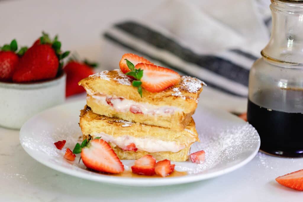 strawberry stuffed French toast cut in half and stacked on top of each other on a white plate. The French toast is topped with strawberries. Maple syrup in a glass jar, a tea towel, and more strawberries are in the background