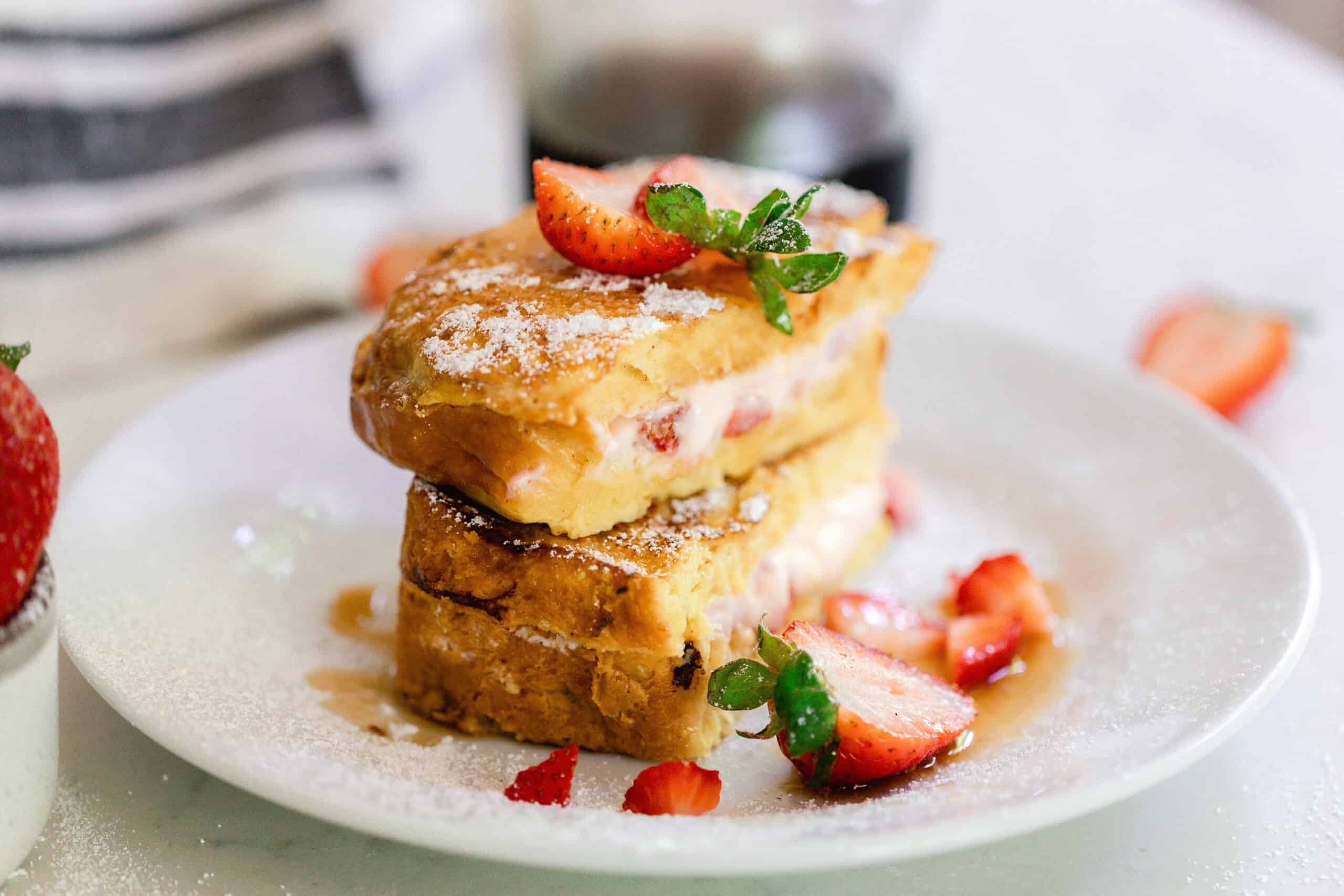 Strawberry stuffed French toast sliced in half and stacked on top of each other topped with powdered sugar and fresh strawberries on a white plate