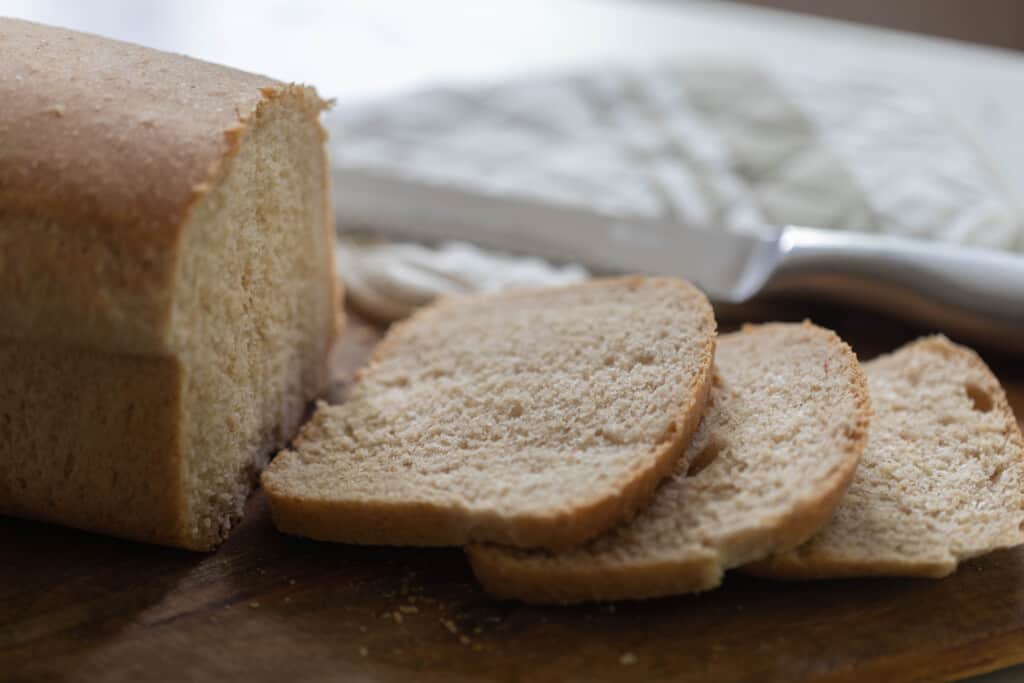 loaf of sourdough whole wheat sandwich bread with three slices cut from the loaf and laying on a cutting board with a towel in the background