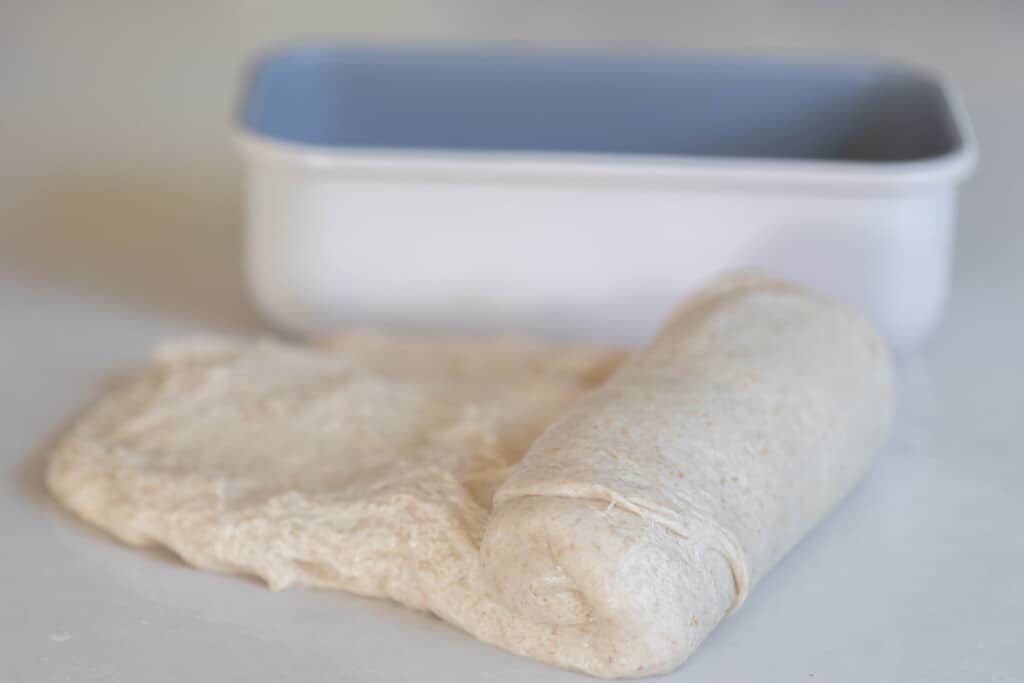 rolling up bread dough into a log on a white quartz countertop with a white loaf pan in the background 