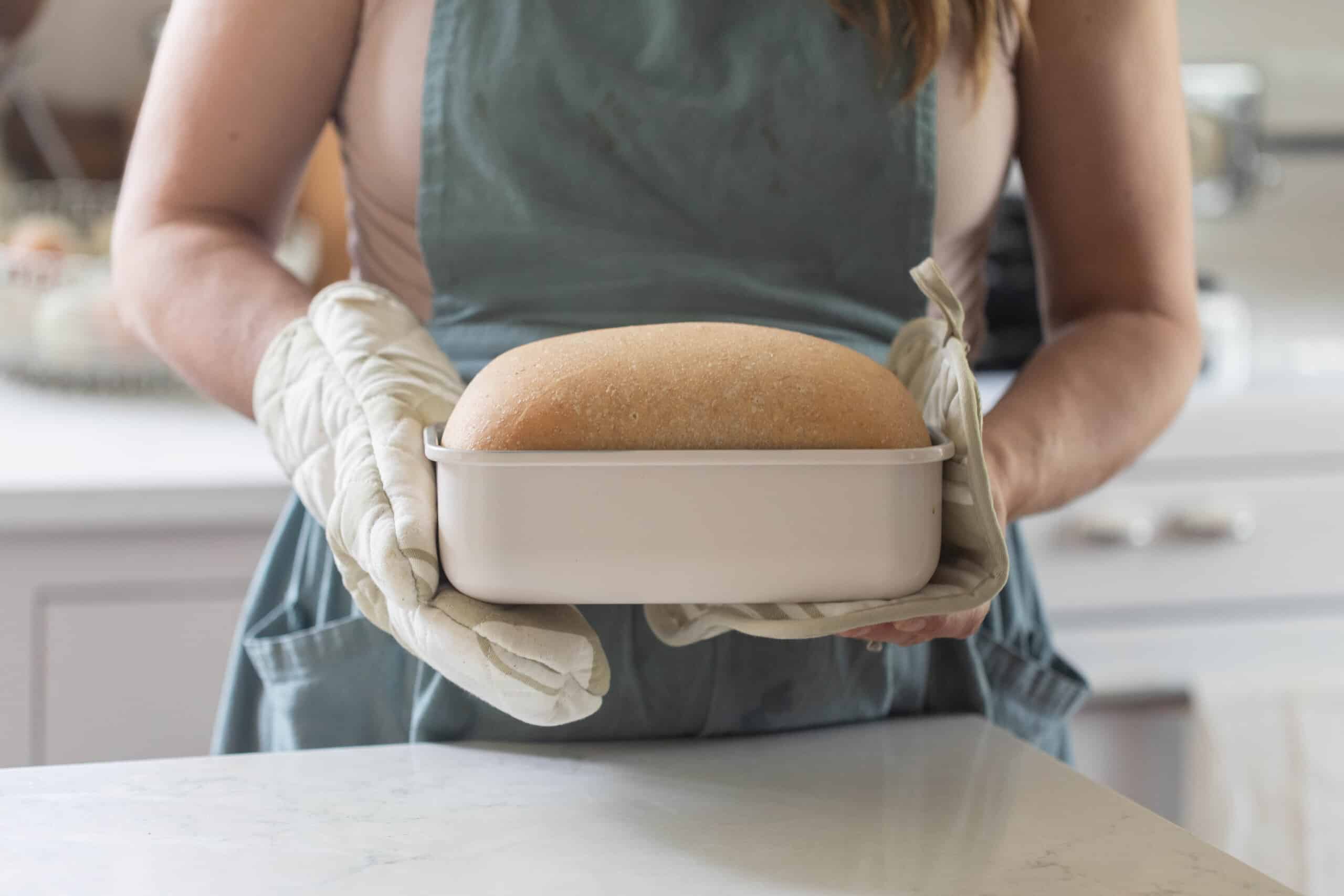 woman wearing a blue apron holding a white loaf pan of bread