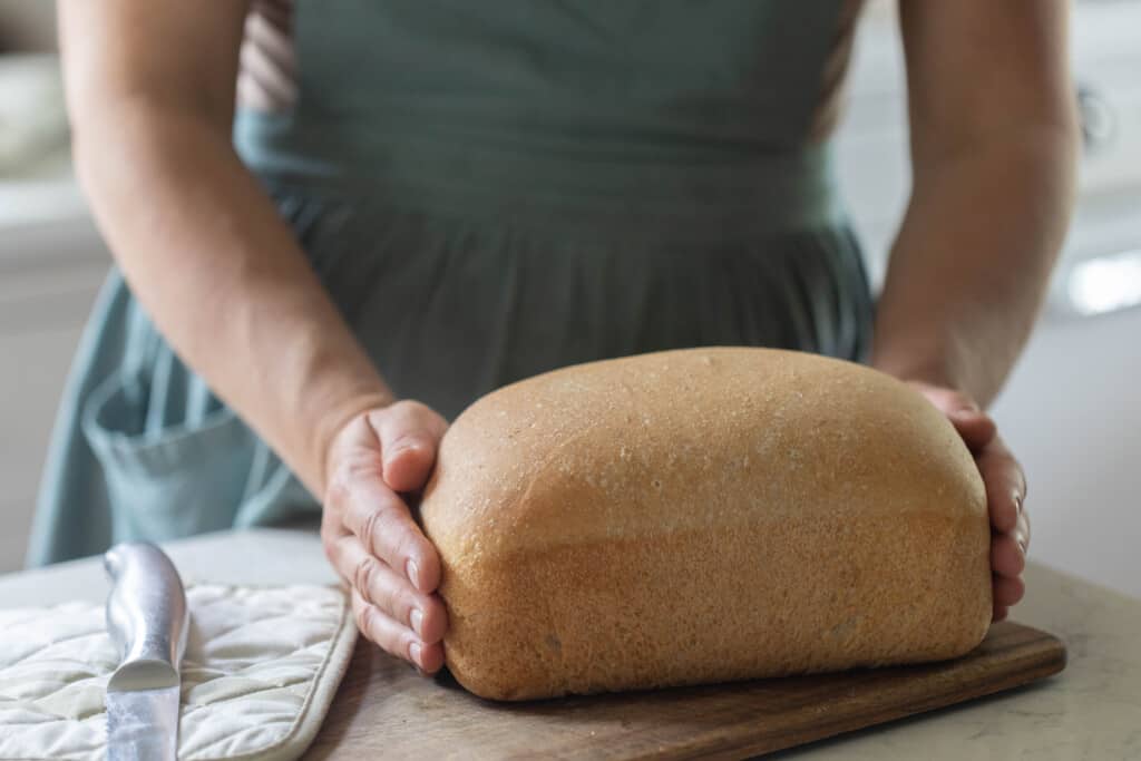 woman wearing a green apron holding. a loaf of whole wheat sourdough sandwich bread on a white countertop