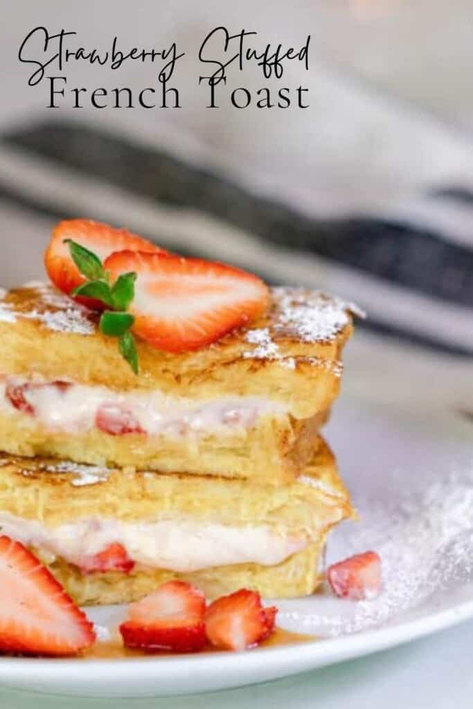 close up of strawberry stuffed French toast cut in half and stacked two high on a white plate. The French toast are topped with halved strawberries and more strawberries are scattered around the plate