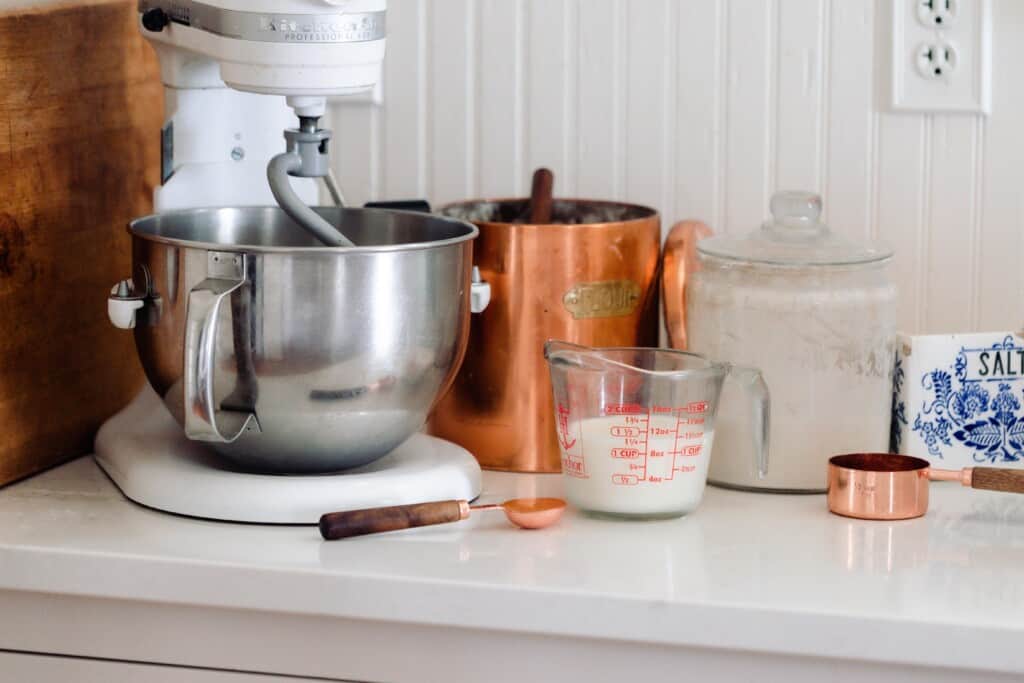 ingredients in a stand mixer bowl with a dough hook attachment on a white countertop with a copper canister to the right