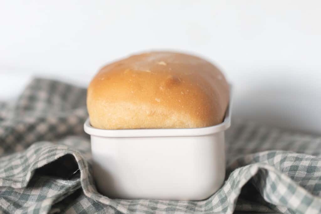 loaf of butter sourdough bread in a white loaf pan on top of a green and white checked towel