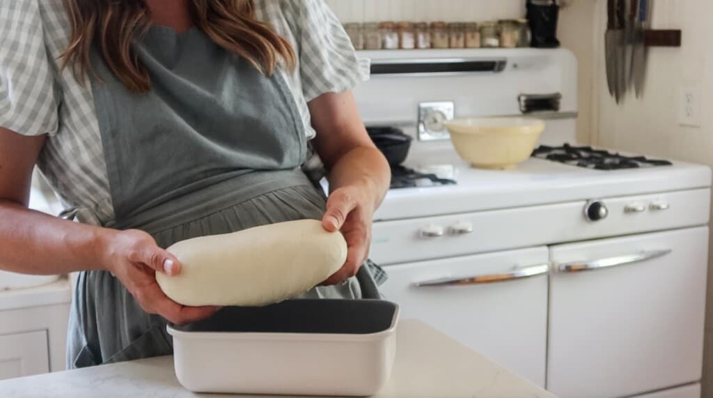 woman wearing a green apron adding bread dough to a loaf pan