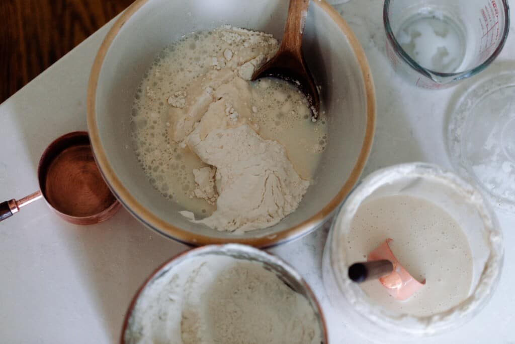 mixing flour, buttermilk, and sourdough starter in a large bowl with canisters of the ingredients surrounding the bowl