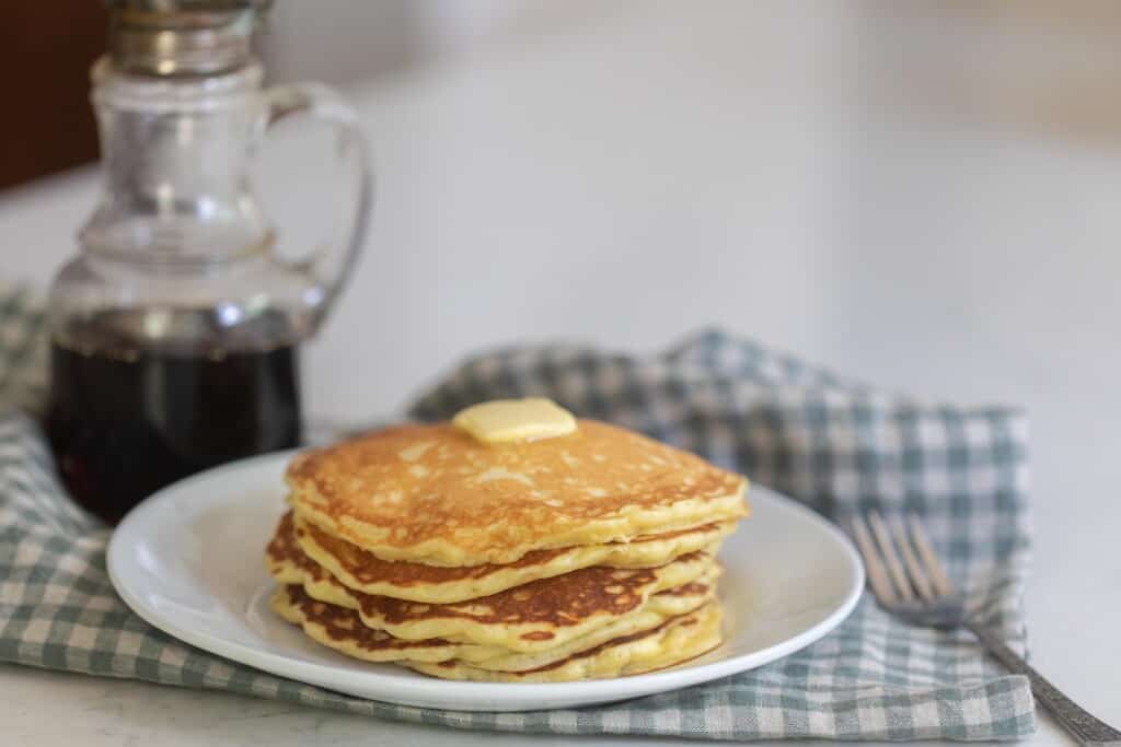 a white plate filled with a stack of sourdough buttermilk pancakes topped with a pat of butter on a white and blue checked plate on a white countertop with a jar of maple syrup in the background