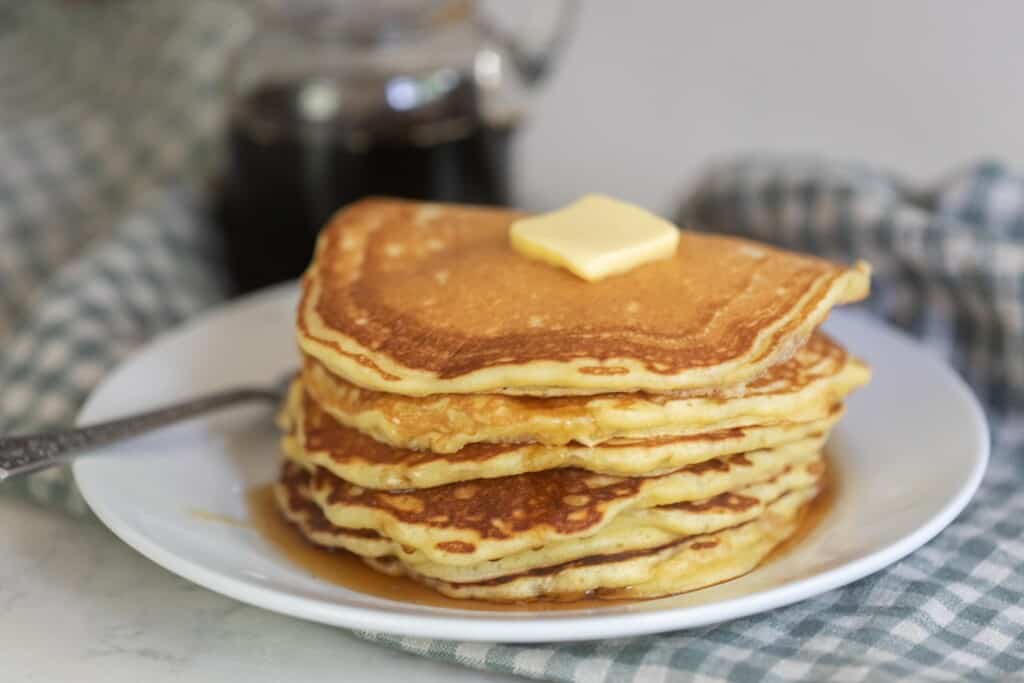 a stack of sourdough buttermilk pancakes on white plate. The pancakes are topped with butter ad there is a jar of maple syrup in the background