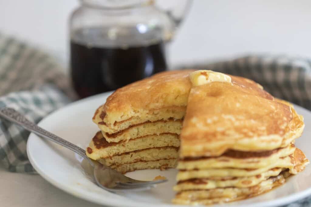 a white plate with a stack of sourdough buttermilk pancake topped with butter and a slice taken out. The plate sits on white and blue checked towel