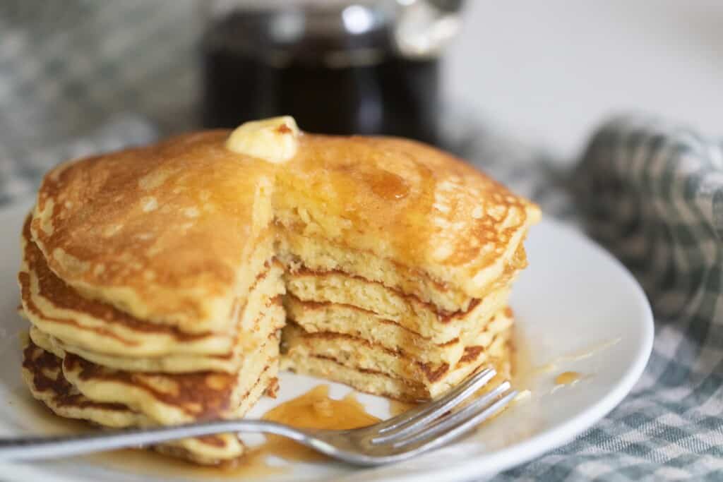 stack of sourdough buttermilk pancakes with a portion cut out. The pancakes are placed on a white plate on a blue and white checked towel with maple syrup in the background