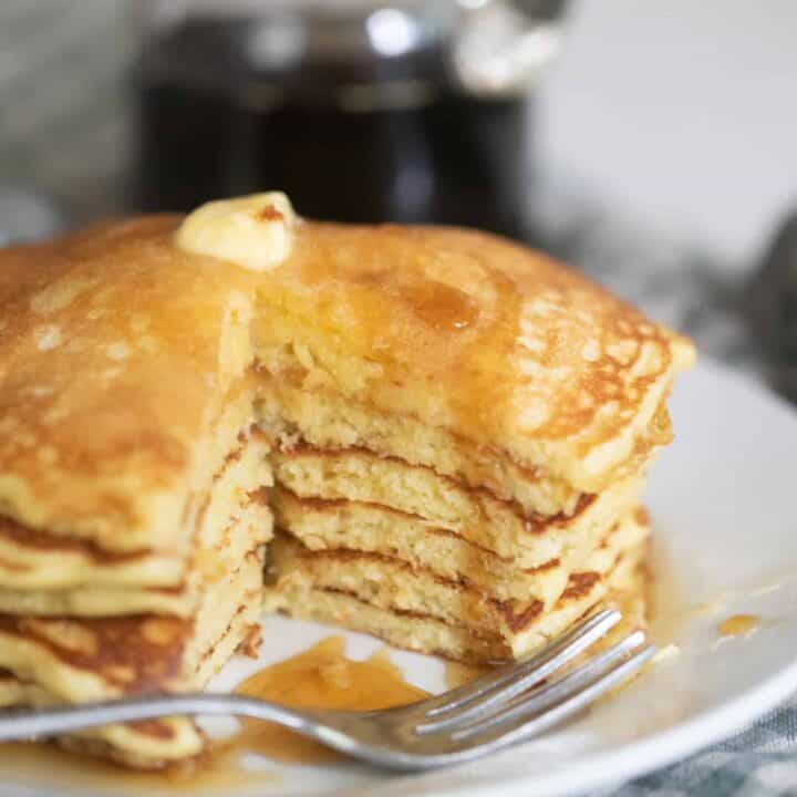 stack of sourdough buttermilk pancakes with a portion cut out. The pancakes are placed on a white plate on a blue and white checked towel with maple syrup in the background