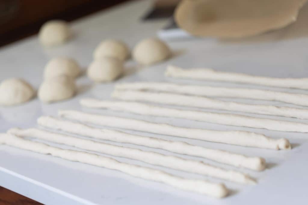 discard pretzel dough rolled into ropes on a white countertop with more dough balls to the left