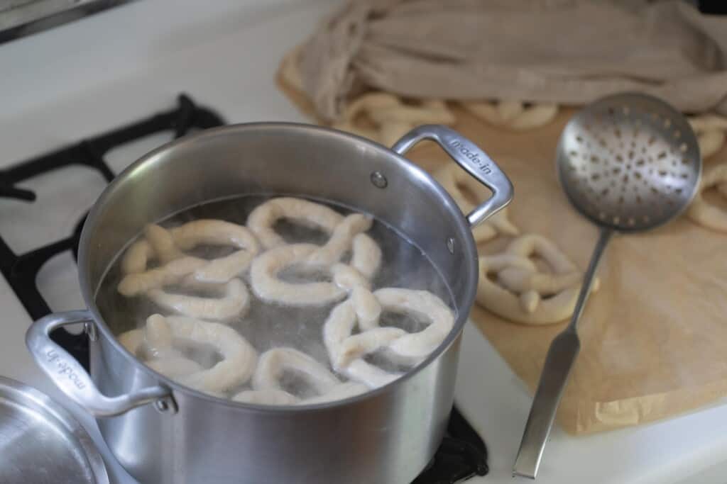 boiling pretzels in baking soda and brown sugar water with more pretzels to the right on parchment paper
