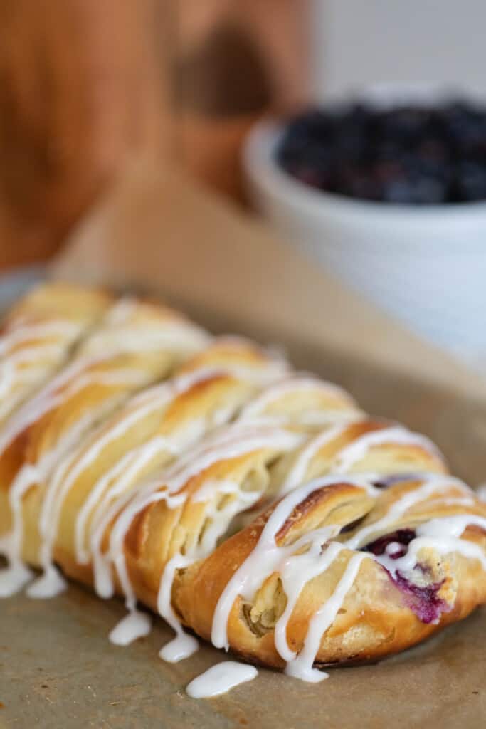 sourdough pastry braid with blueberry cream cheese filling drizzled with icing on parchment paper with a bowl of blueberries in the background
