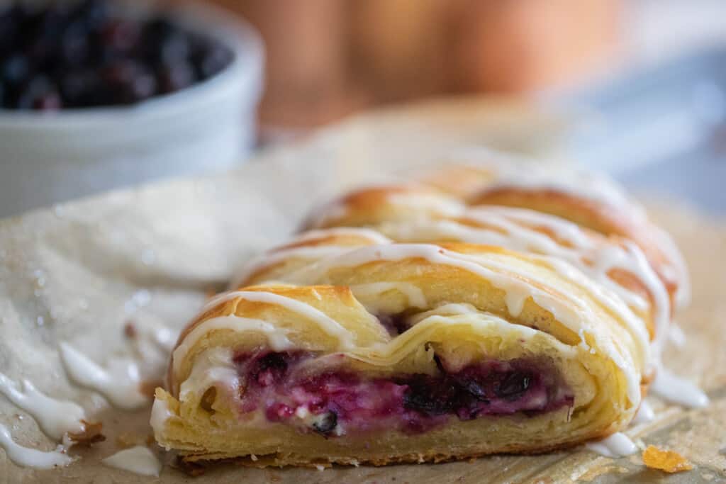 sourdough pastry braid with a slice off exposing the blueberry cream cheese filling on parchment paper with a bowl of blueberries in the background