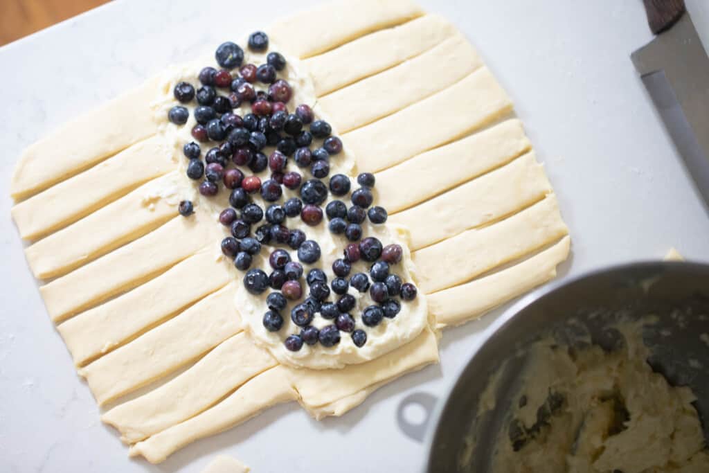pastry dough with slits cut along the sides and cream cheese filling topped with blueberries down the middle