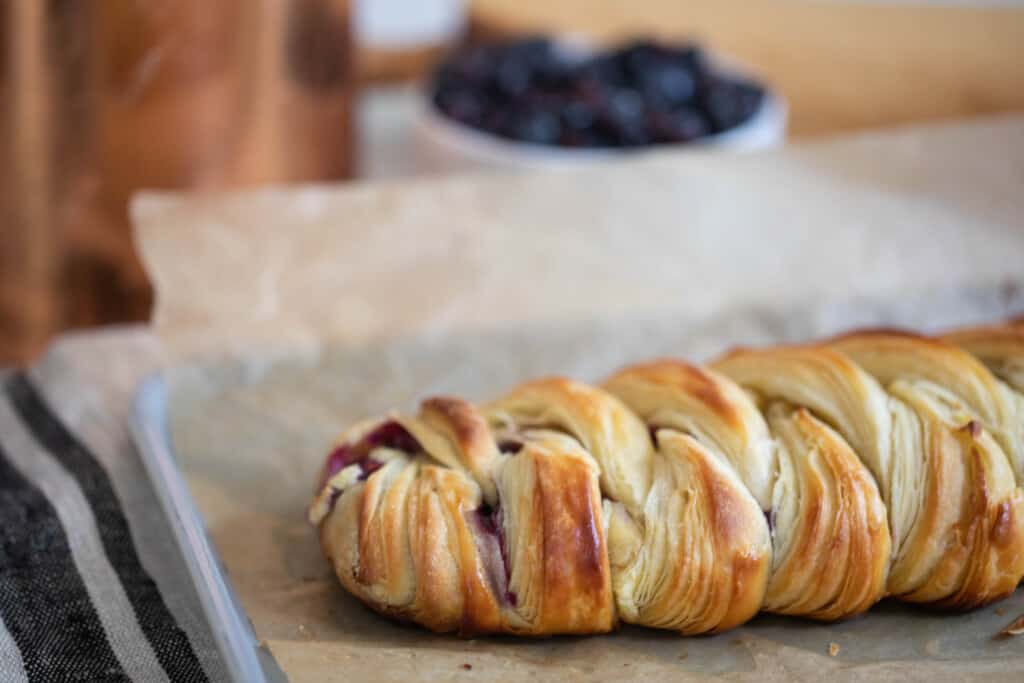 sourdough pastry braid on a parchment lined baking sheet with a copper container and a bowl of blueberries in the background