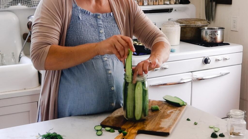 woman wearing a blue dress adding sliced cucumbers to a jar. The jar is on a wood cutting board on a white countertop 