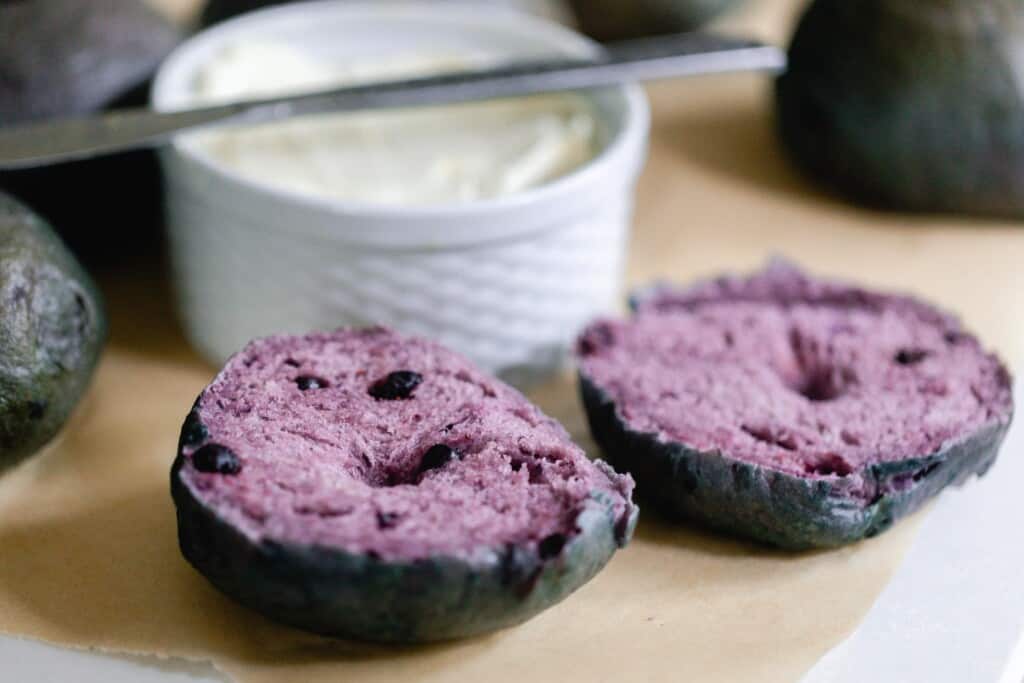 sourdough blueberry bagel sliced in half on parchment paper with a container of cream cheese behind the bagels