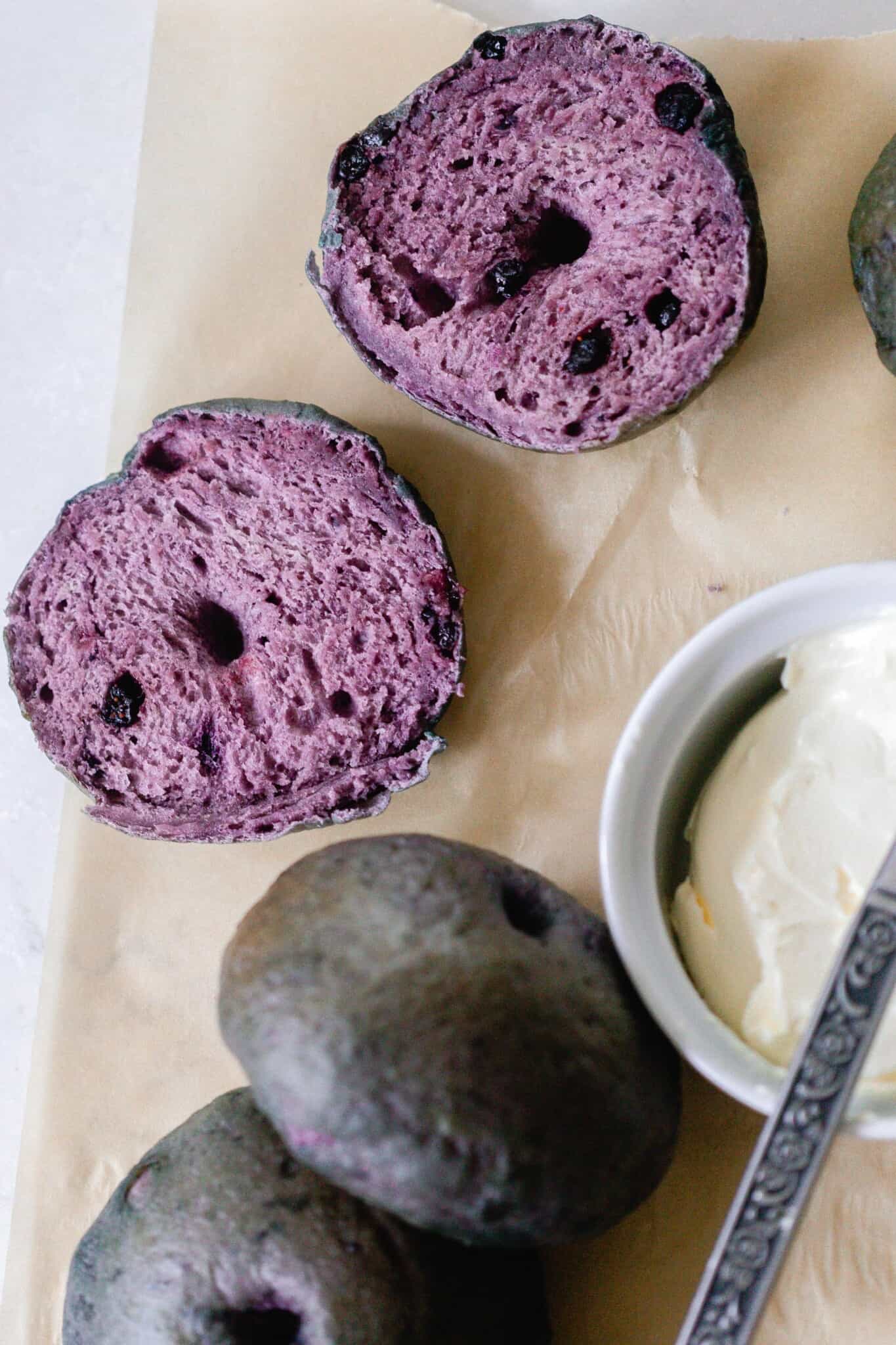 two blueberry sourdough bagels and one bagel sliced in half on parchment paper with a container of cream cheese to the left